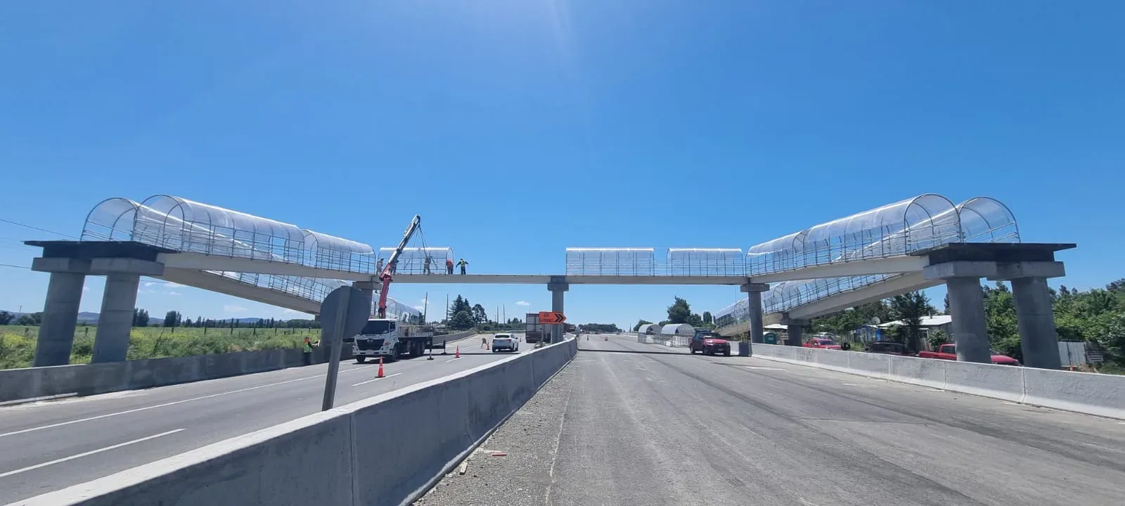 Camión pluma montando cúpulas sobre las dos pasarelas peatonales que cruzan la carretera, visto desde la mediana entre calzadas